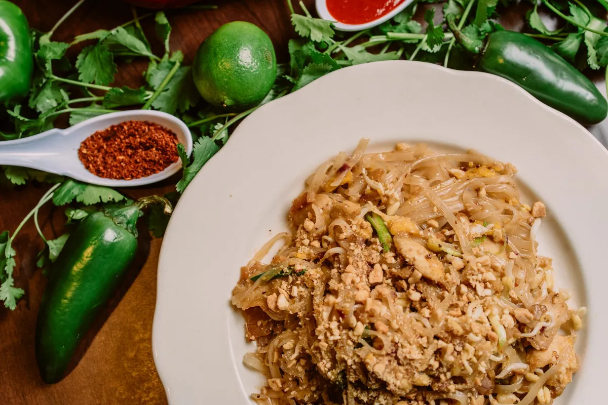 Pad Thai on a white plate with ground peanuts, surrounded by peppers and lime.