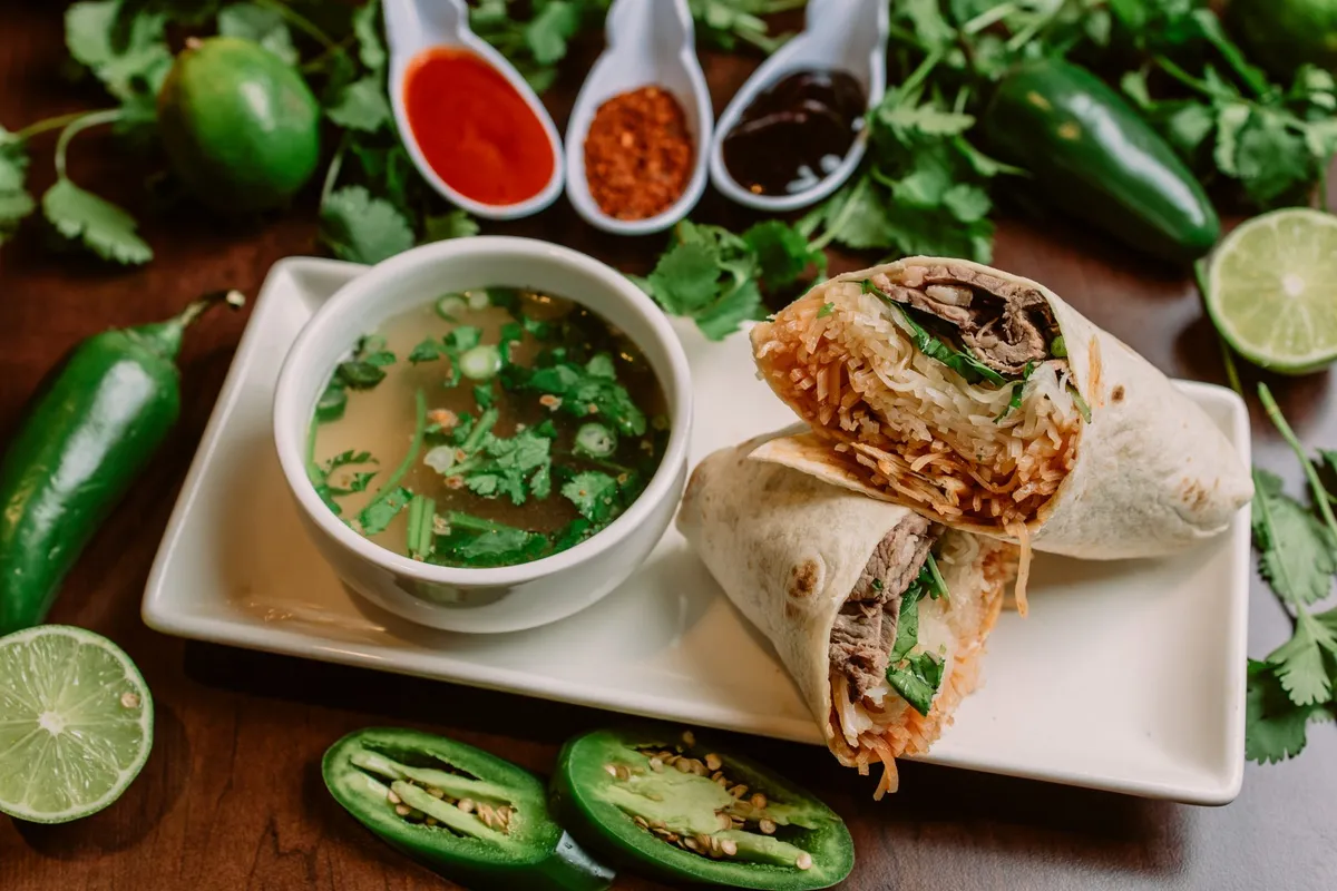 A sliced wrap served with a cup of broth, lime and chili sauces.